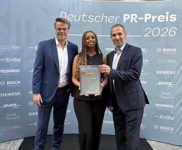 Drei Personen präsentieren eine Bronze-Auszeichnung beim Deutschen PR-Preis 2026 vor einer Veranstaltungswand mit Sponsorenlogos