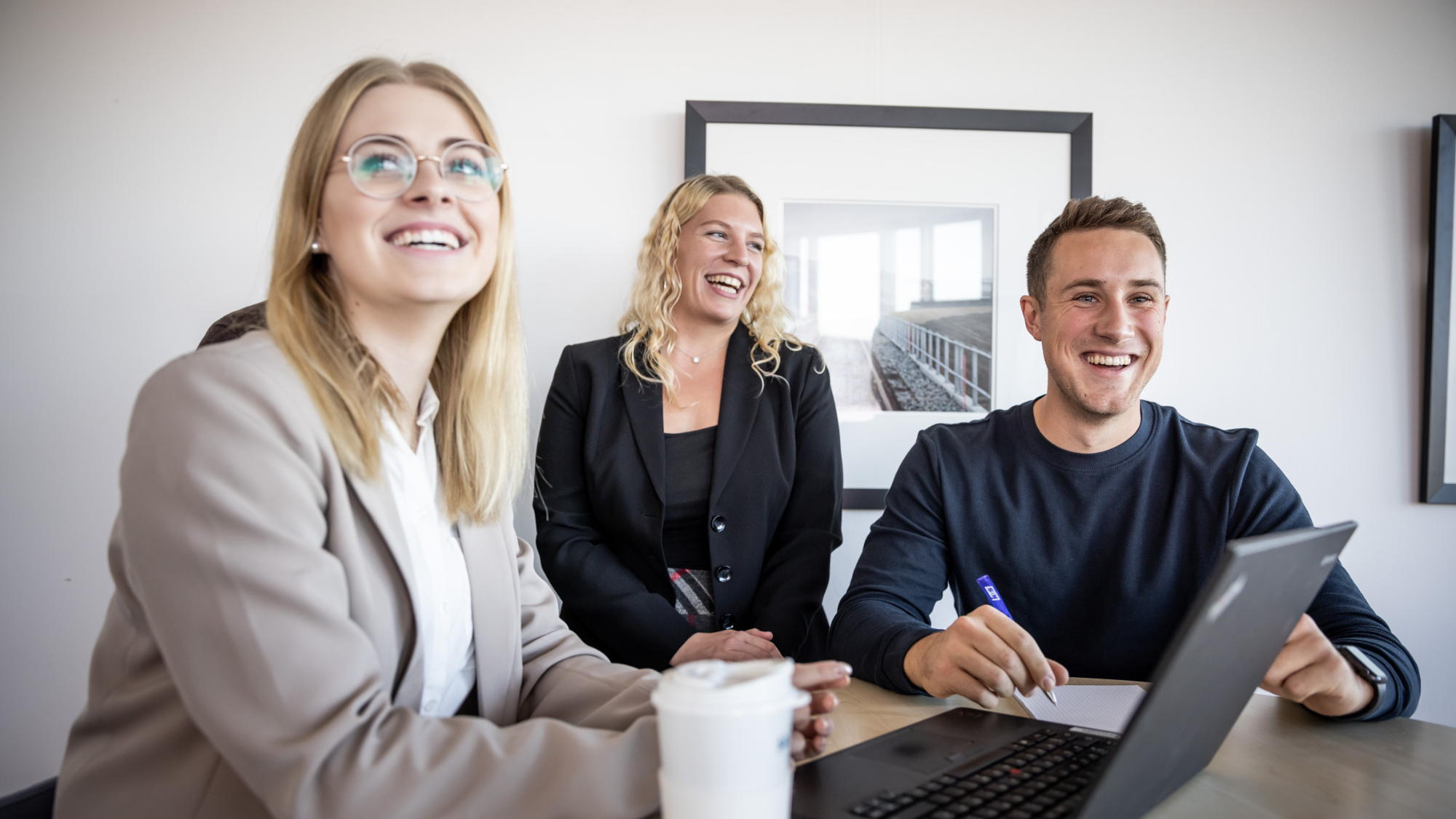 Drei Studierende sitzen gemeinsam am Tisch, lachen und arbeiten mit Laptop und Notizen.