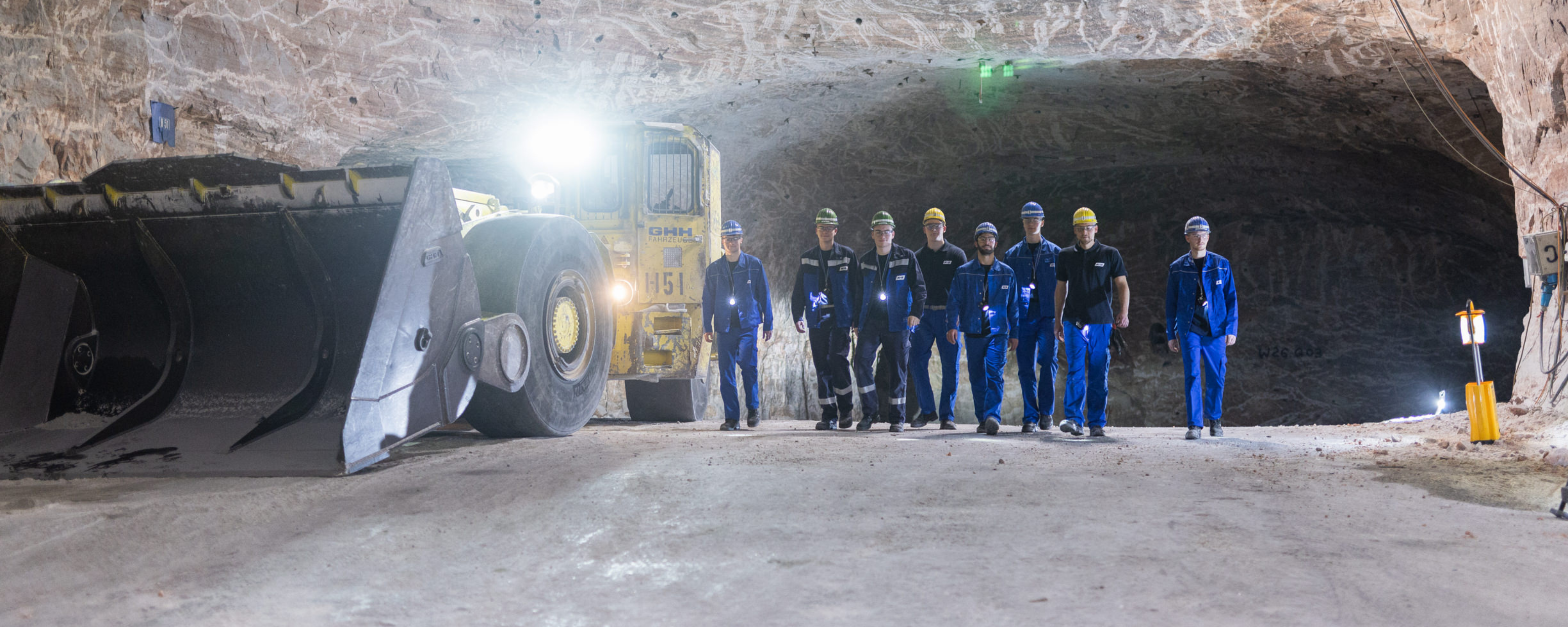 Mitarbeitende gehen unter Tage auf die Kamera zu, begleitet von einem großen Lader.
