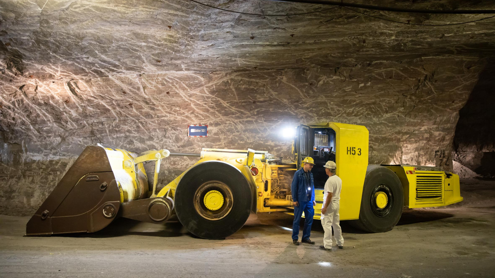 Zwei K+S Mitarbeitende unter Tage im Gespräch neben einem gelben Bergbaufahrzeug.