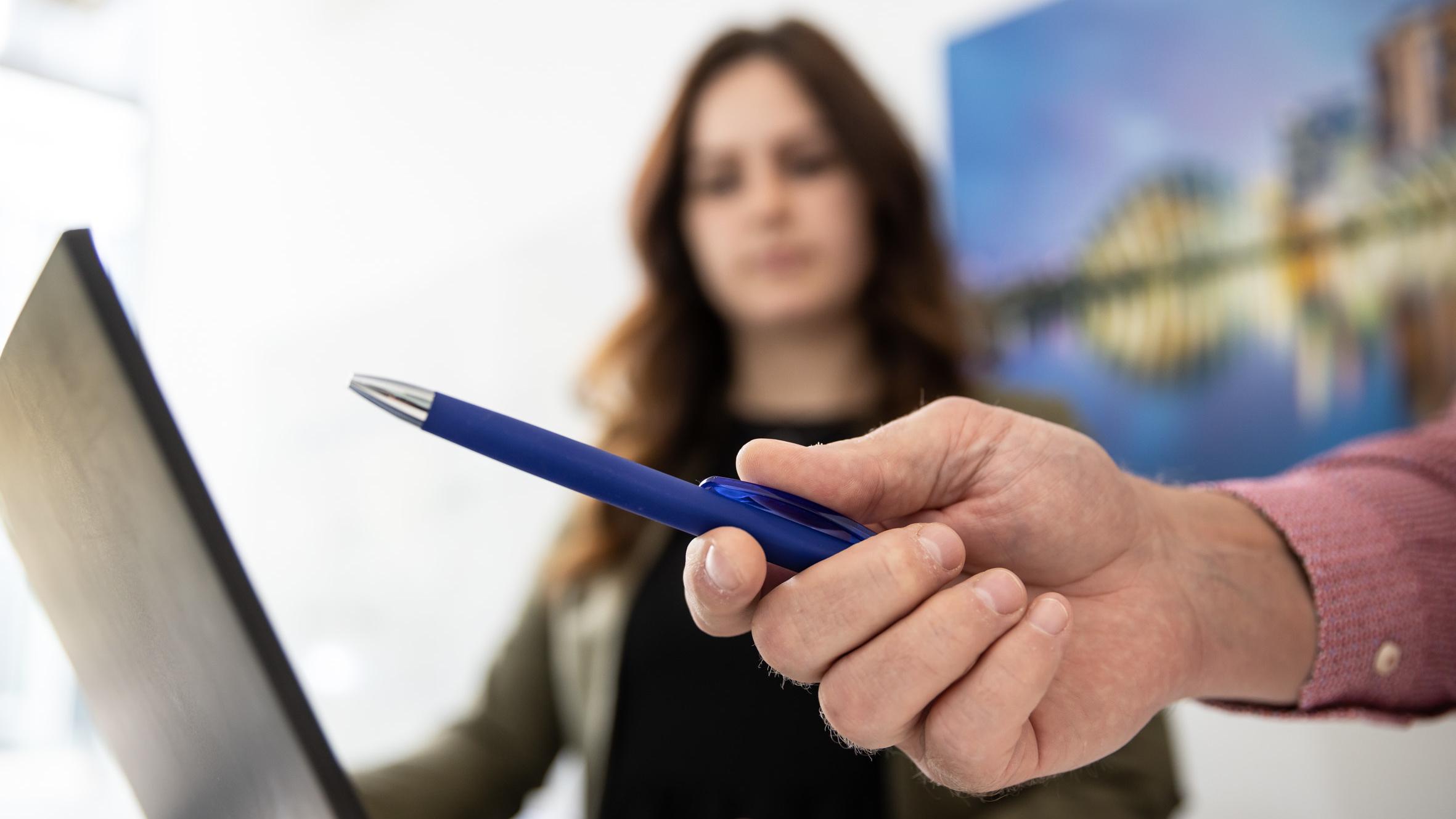 Close-up of a hand holding a blue ballpoint pen, with a person in the background in front of a laptop.