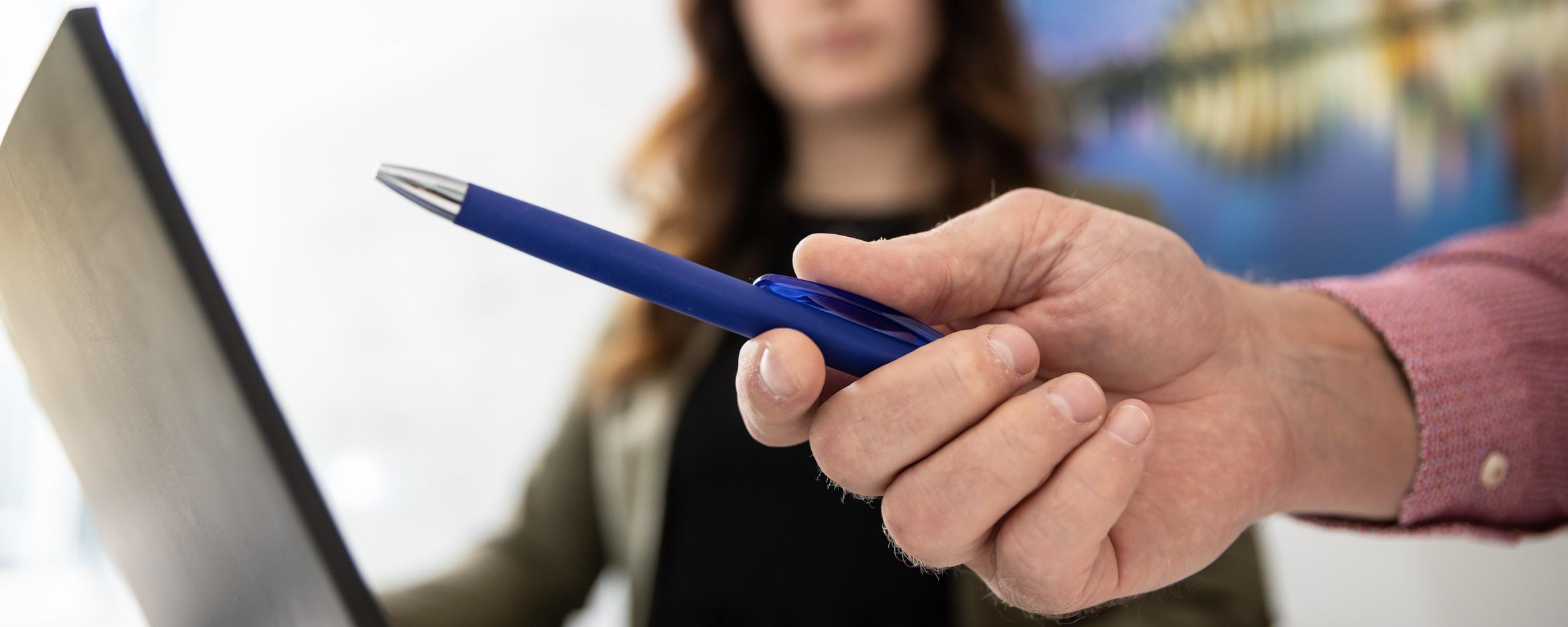 Close-up of a hand holding a blue ballpoint pen, with a person in the background in front of a laptop.