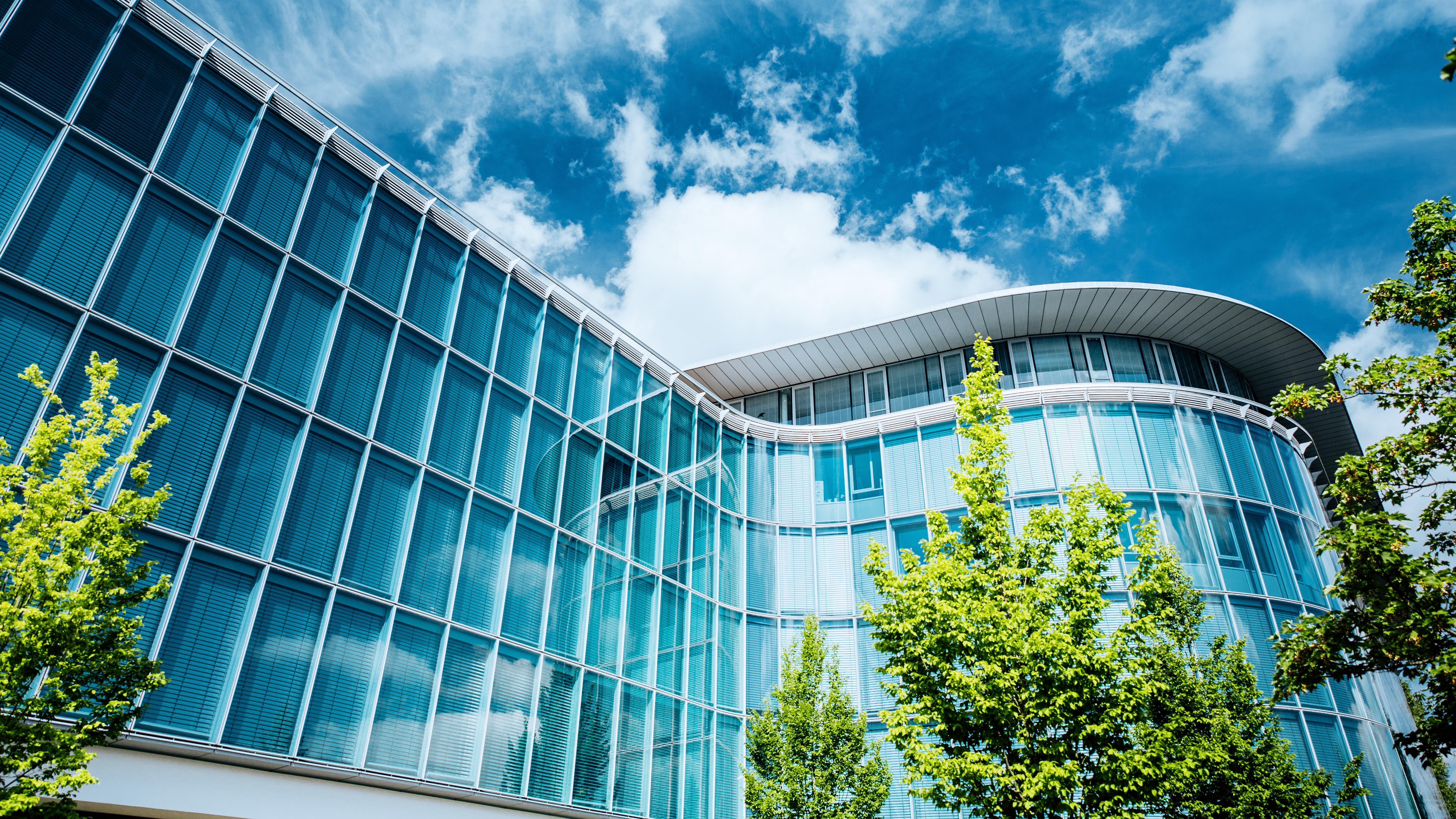 Modern glass façade of an office building with a curved roof, green trees in front, under a blue sky with white clouds.