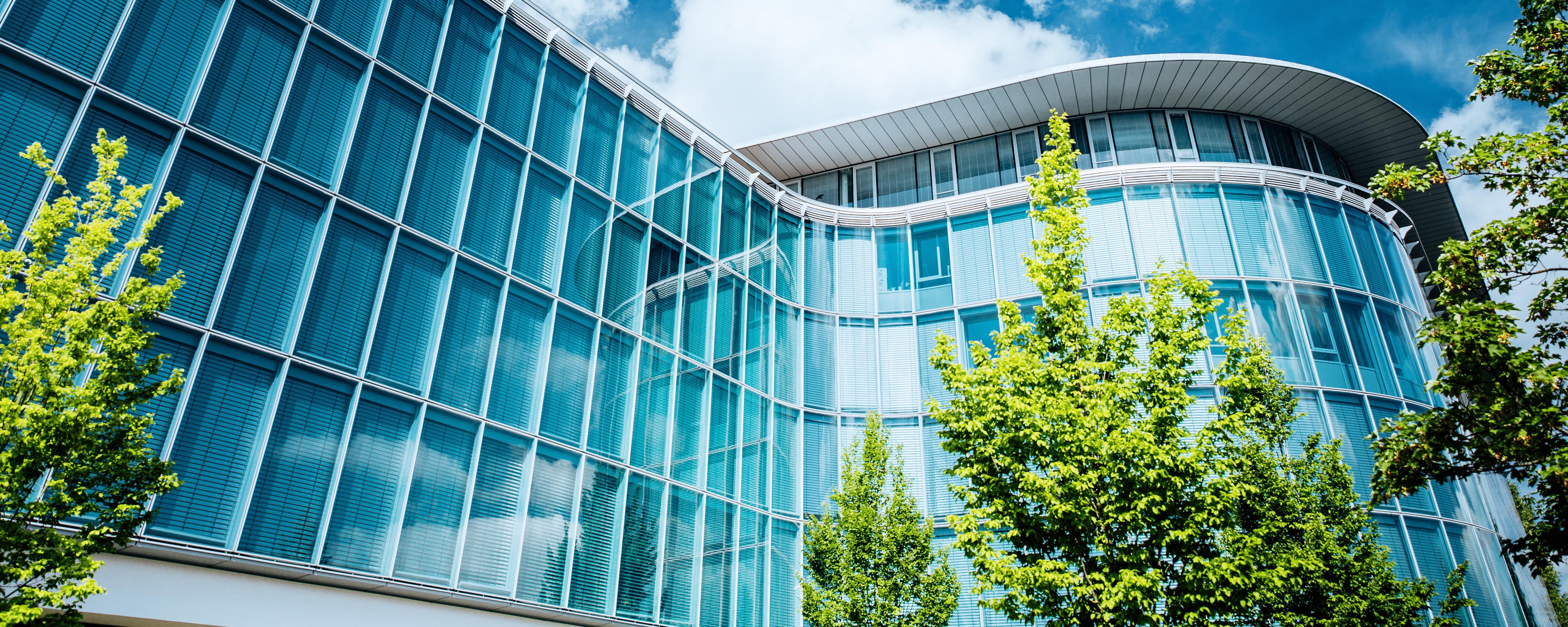 Modern glass façade of an office building with a curved roof, green trees in front, under a blue sky with white clouds.