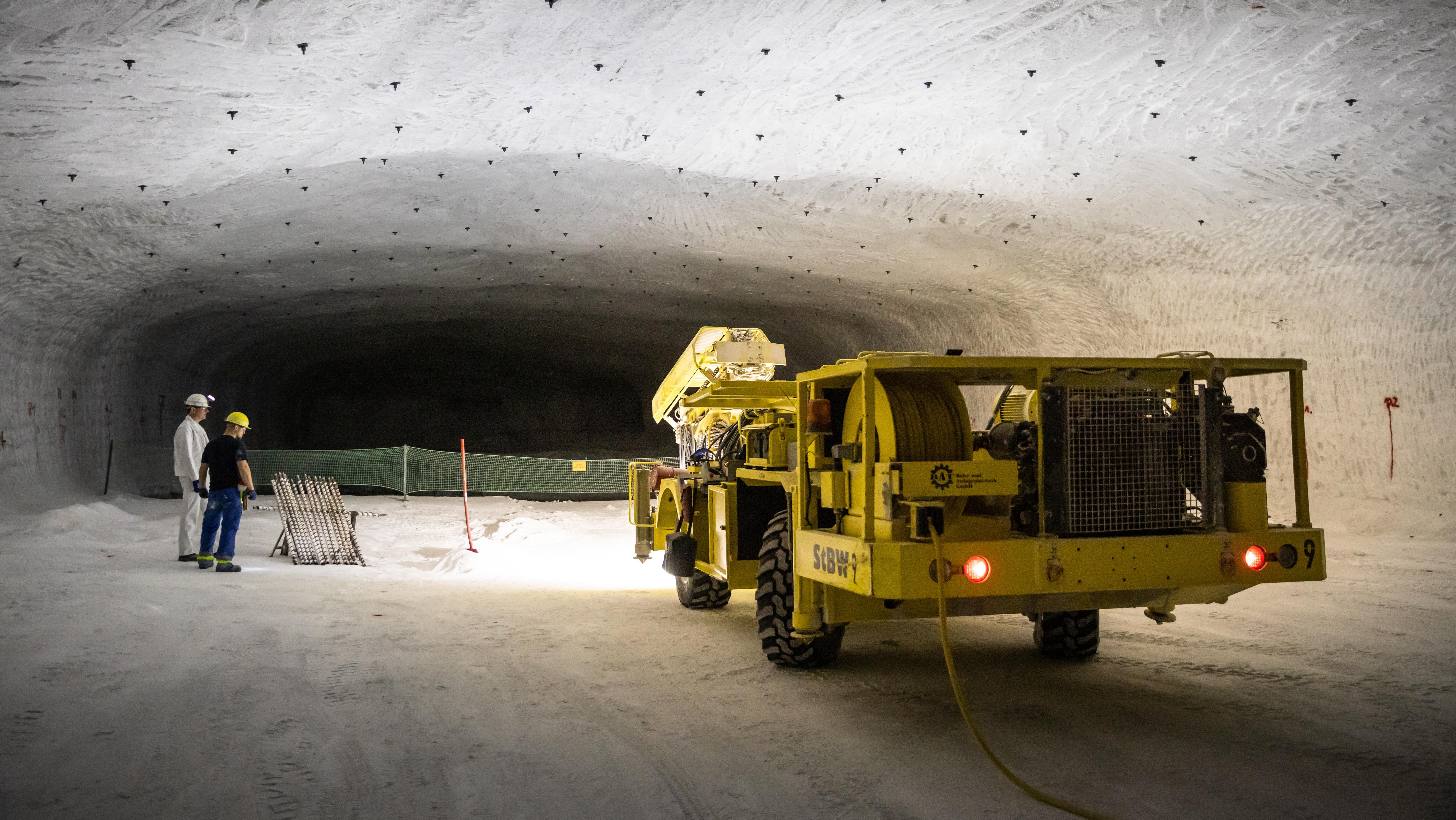 Underground mining area with two workers in protective clothing standing next to a yellow specialized vehicle in a large salt tunnel.