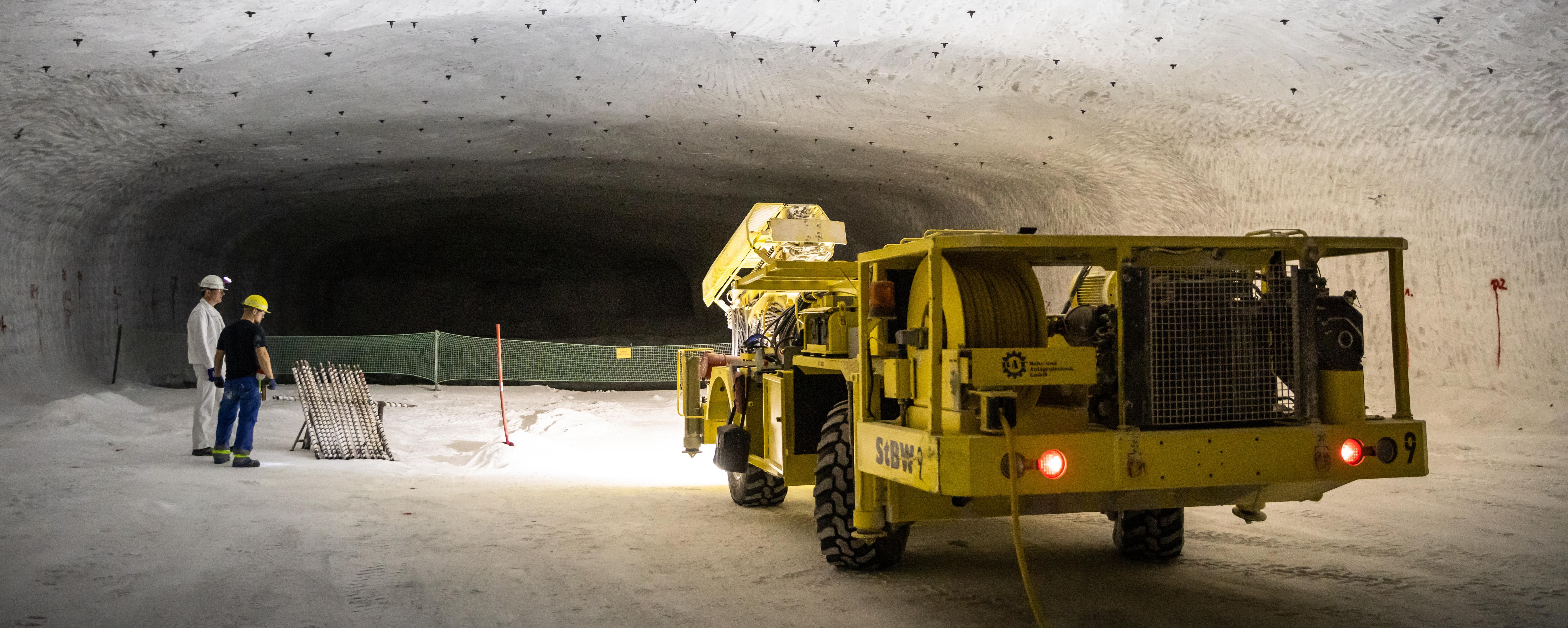 Underground mining area with two workers in protective clothing standing next to a yellow specialized vehicle in a large salt tunnel.