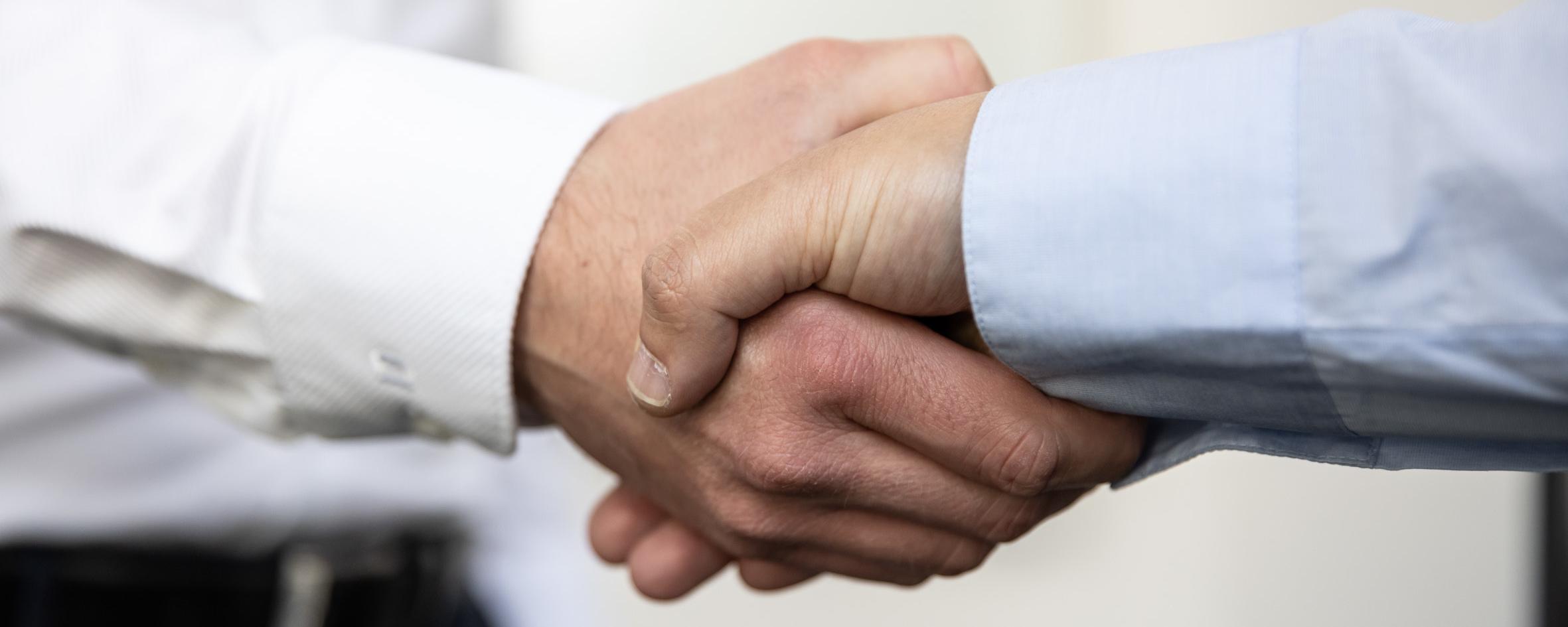 Close-up of a handshake between two people. Both are wearing long-sleeved shirts, one white and the other light blue. The background is blurred and bright, keeping the focus on the hands.