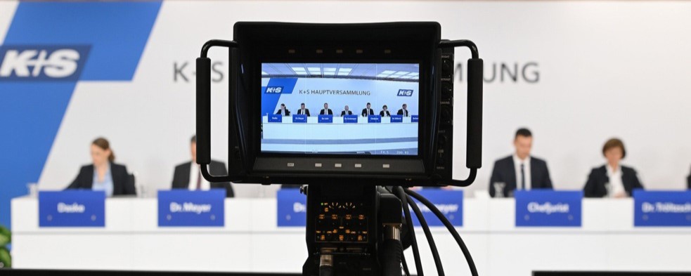 Eine Kamera steht im Vordergrund und filmt eine Podiumsdiskussion. Auf dem Kameradisplay ist eine Nahaufnahme des Podiums mit mehreren Personen und dem Text „K+S Hauptversammlung“ zu sehen. Im Hintergrund befindet sich eine weiße Wand mit dem K+S-Logo und dem Schriftzug „Hauptversammlung“. Auf dem Tisch vor den Personen sind blaue Namensschilder sichtbar.