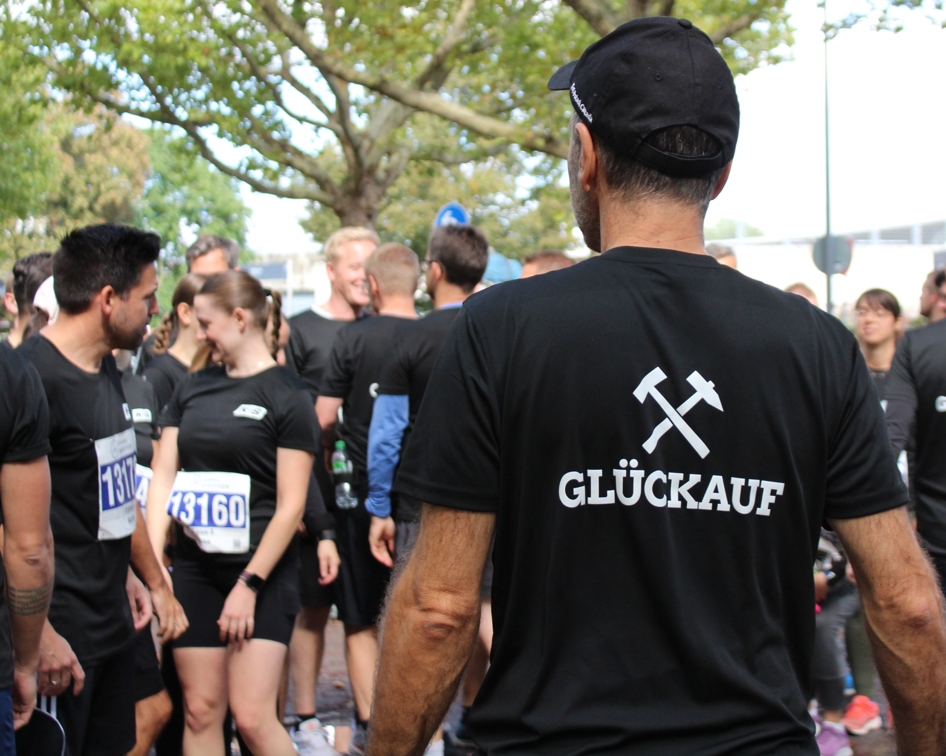 Eine Gruppe von Menschen nimmt an einem Outdoor-Event teil, vermutlich einem Firmenlauf. Die Teilnehmenden tragen schwarze T-Shirts, einige mit Startnummern. Im Vordergrund ist eine Person zu sehen, auf deren Rücken „GLÜCKAUF“ steht, begleitet von zwei gekreuzten Symbolen – Schlägel und Eisen, traditionell aus dem Bergbau. Im Hintergrund sind Bäume und weitere Teilnehmende zu erkennen.