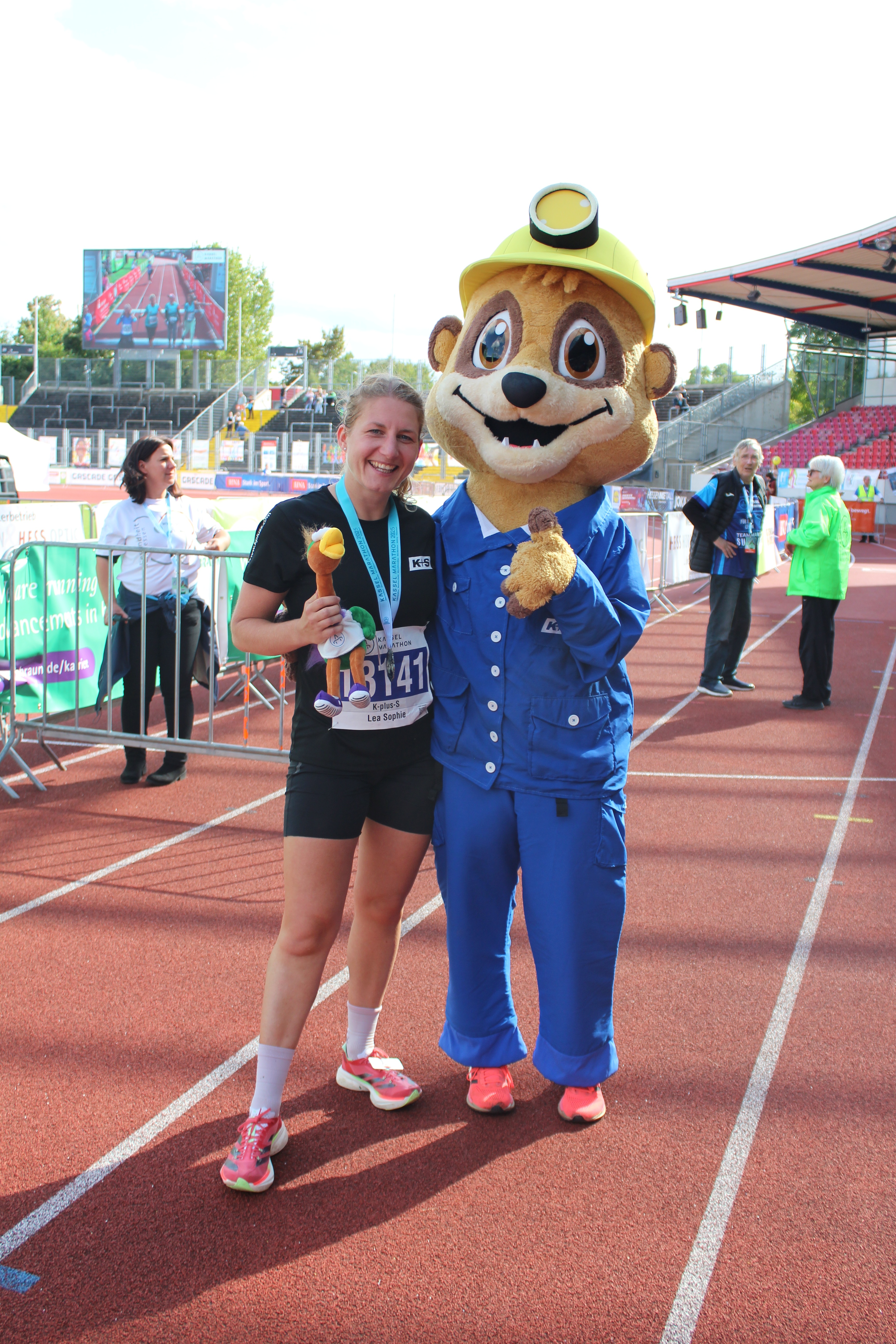 Eine Person steht auf einer Laufbahn und posiert mit dem Maskottchen Salty, das ein blaues Kostüm und einen gelben Helm trägt. Die Person trägt Sportkleidung, darunter pinke Schuhe. In der Hand hält sie einen orangefarbenen Gegenstand und trägt eine Medaille um den Hals – ein Hinweis auf die Teilnahme am Firmenlauf. Im Hintergrund sind weitere Personen und Absperrgitter zu sehen.