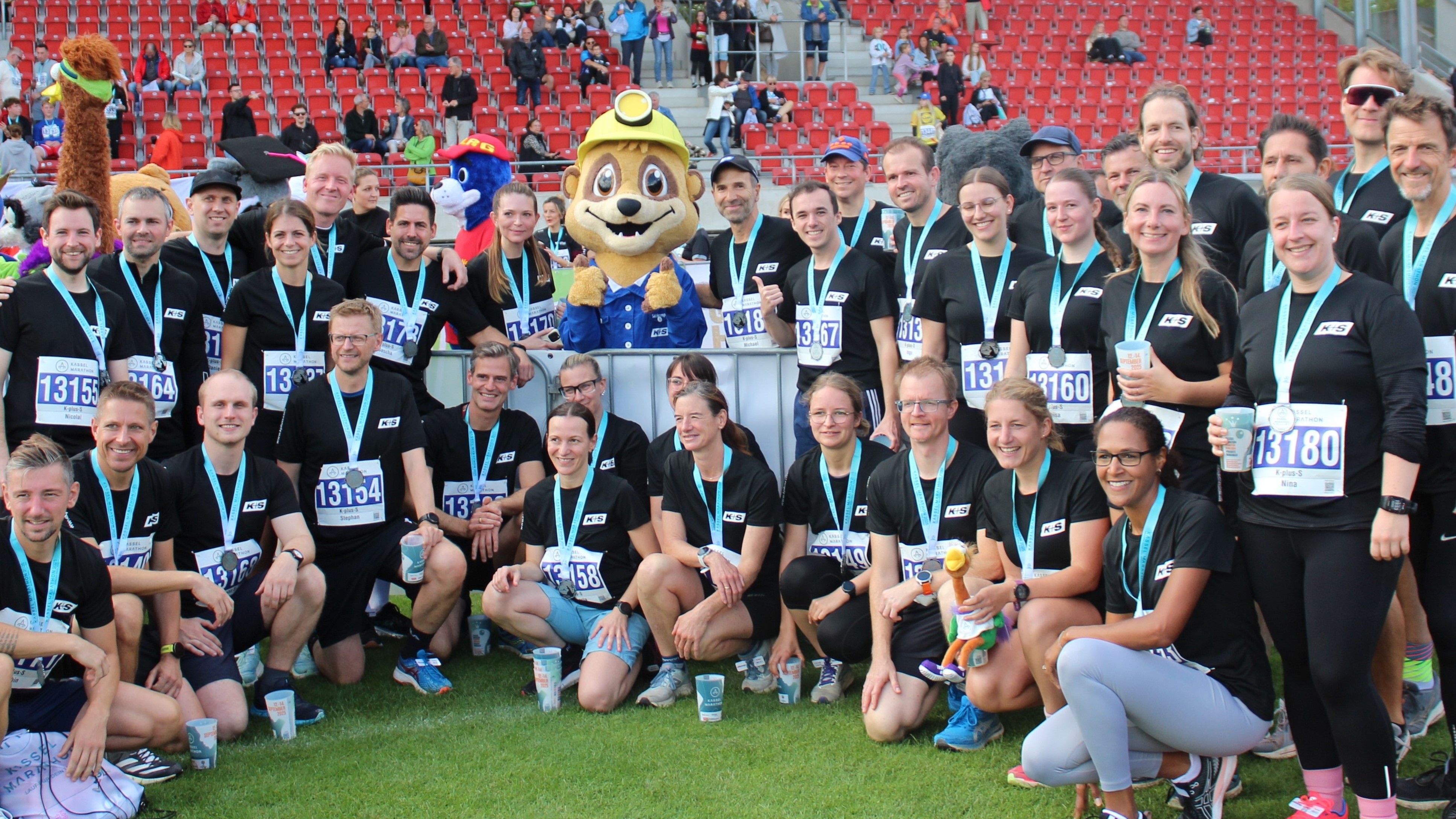 Eine große Gruppe von Menschen in schwarzen T-Shirts und mit Medaillen um den Hals posiert für ein Gruppenfoto auf einem Sportplatz. In der Mitte steht das Maskottchen Salty mit gelbem Helm und blauem Kostüm, das wie eine Zeichentrickfigur aussieht. Im Hintergrund sind rote Tribünen zu sehen.