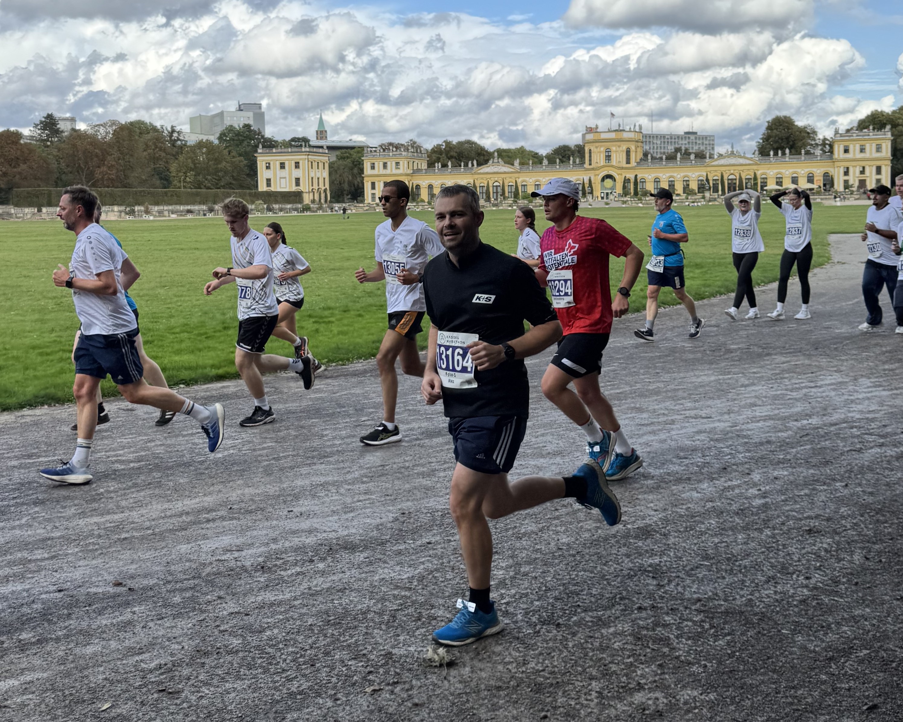 Eine Gruppe von Menschen läuft auf einem Kiesweg im Auepark in Kassel, umgeben von Wiesen und historischen Gebäuden im Hintergrund. Die Läufer tragen Sportkleidung und Startnummern, was auf einen Firmenlauf hinweist. Der Himmel ist teilweise bewölkt, und in der Ferne sind Bäume zu sehen.