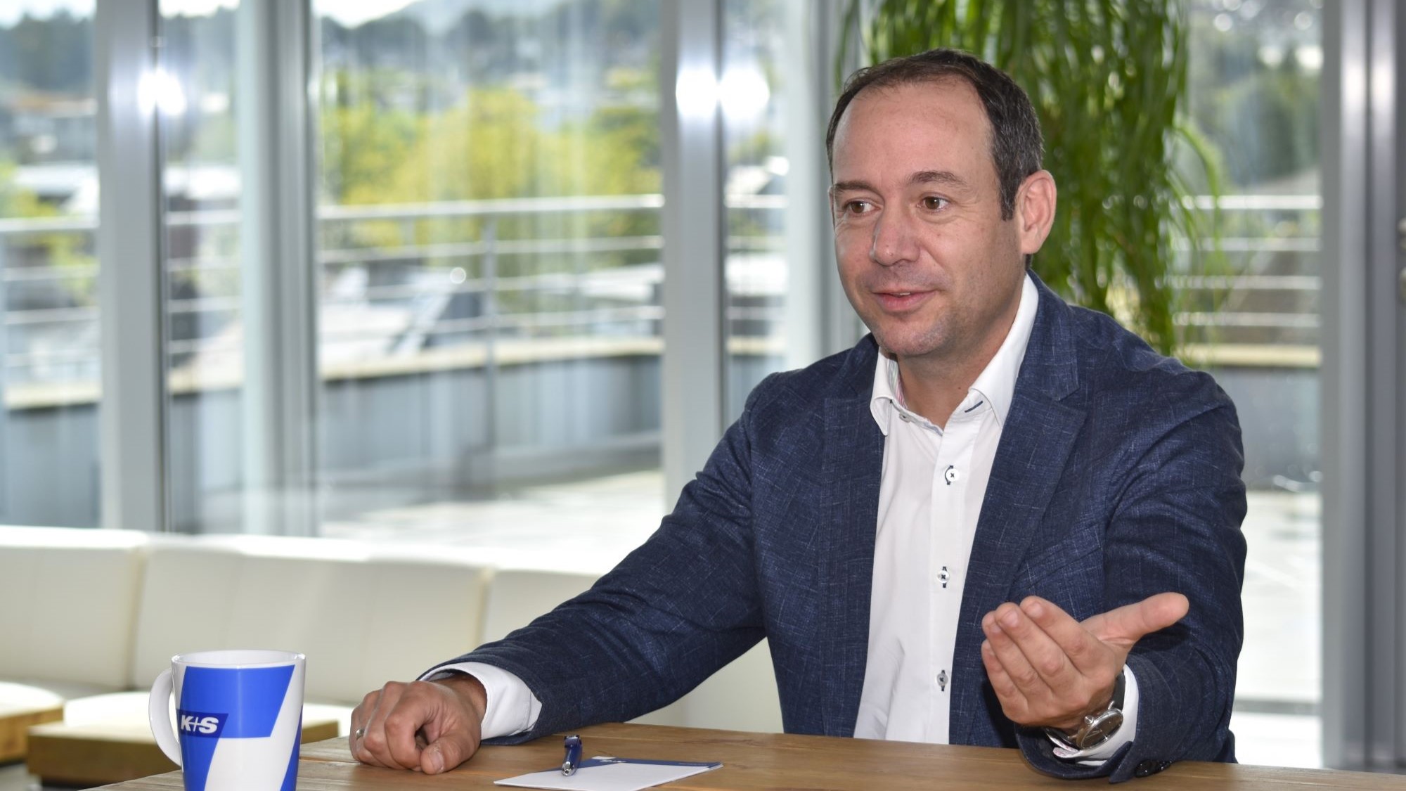 Ein Mann in einem blauen Sakko und weißem Hemd sitzt an einem Holztisch. Vor ihm liegt ein Blatt Papier mit einem Stift darauf. Auf dem Tisch steht eine Tasse mit der Aufschrift „K+S“. Im Hintergrund sind große Fenster, durch die man eine grüne Landschaft und Gebäude sehen kann.