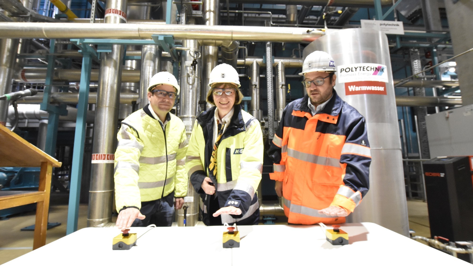 Drei Personen in Schutzkleidung stehen in einer Industriehalle vor einer komplexen Rohrleitungsanlage. Sie berühren jeweils einen Bedienknopf auf einem Tisch vor ihnen.
