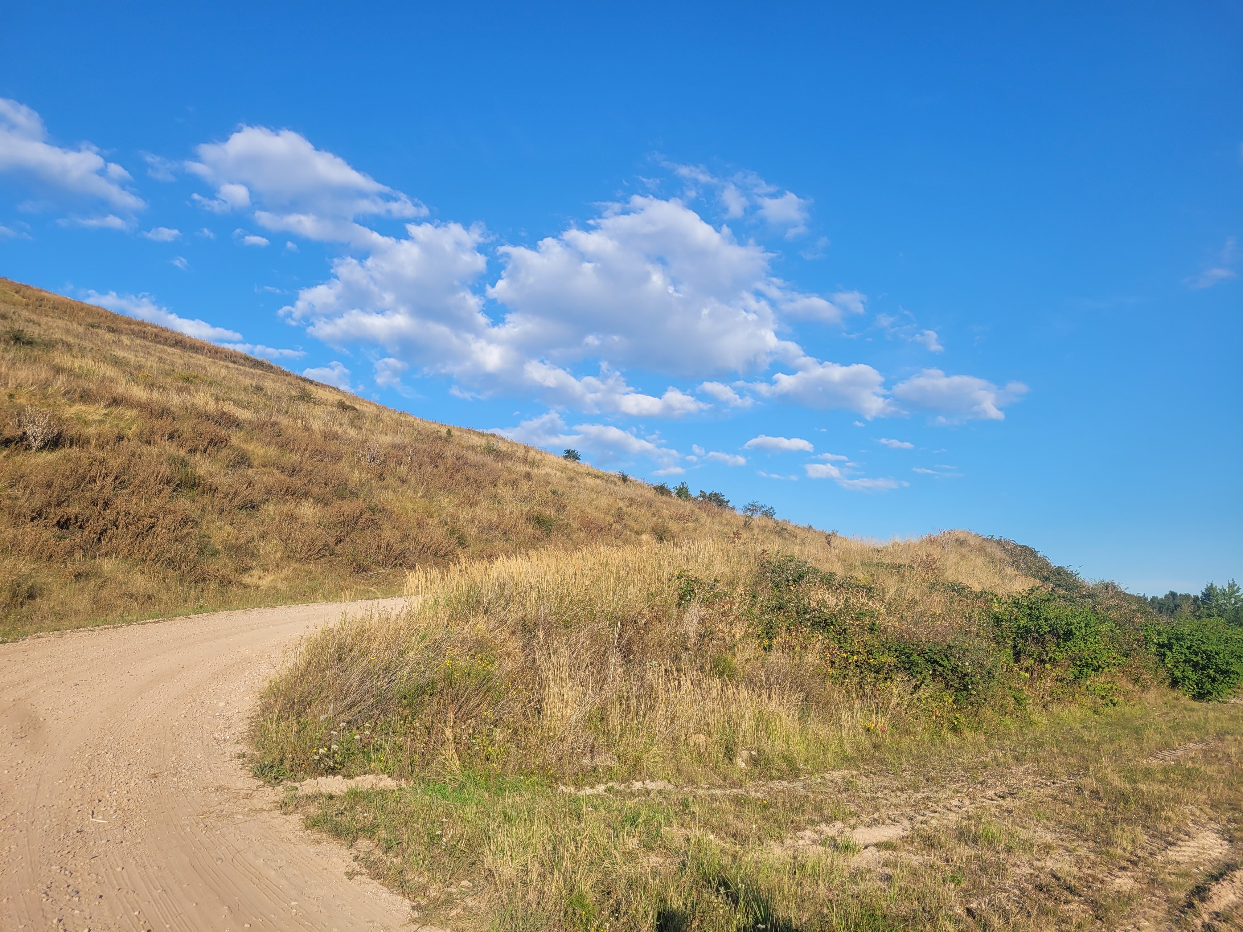 Das Bild zeigt einen unbefestigten Weg, der sich um eine grasbewachsene Halde windet. Der Himmel ist klar und blau mit vereinzelten weißen Wolken. Die Halde ist mit trockenem Gras und einigen grünen Sträuchern bedeckt.