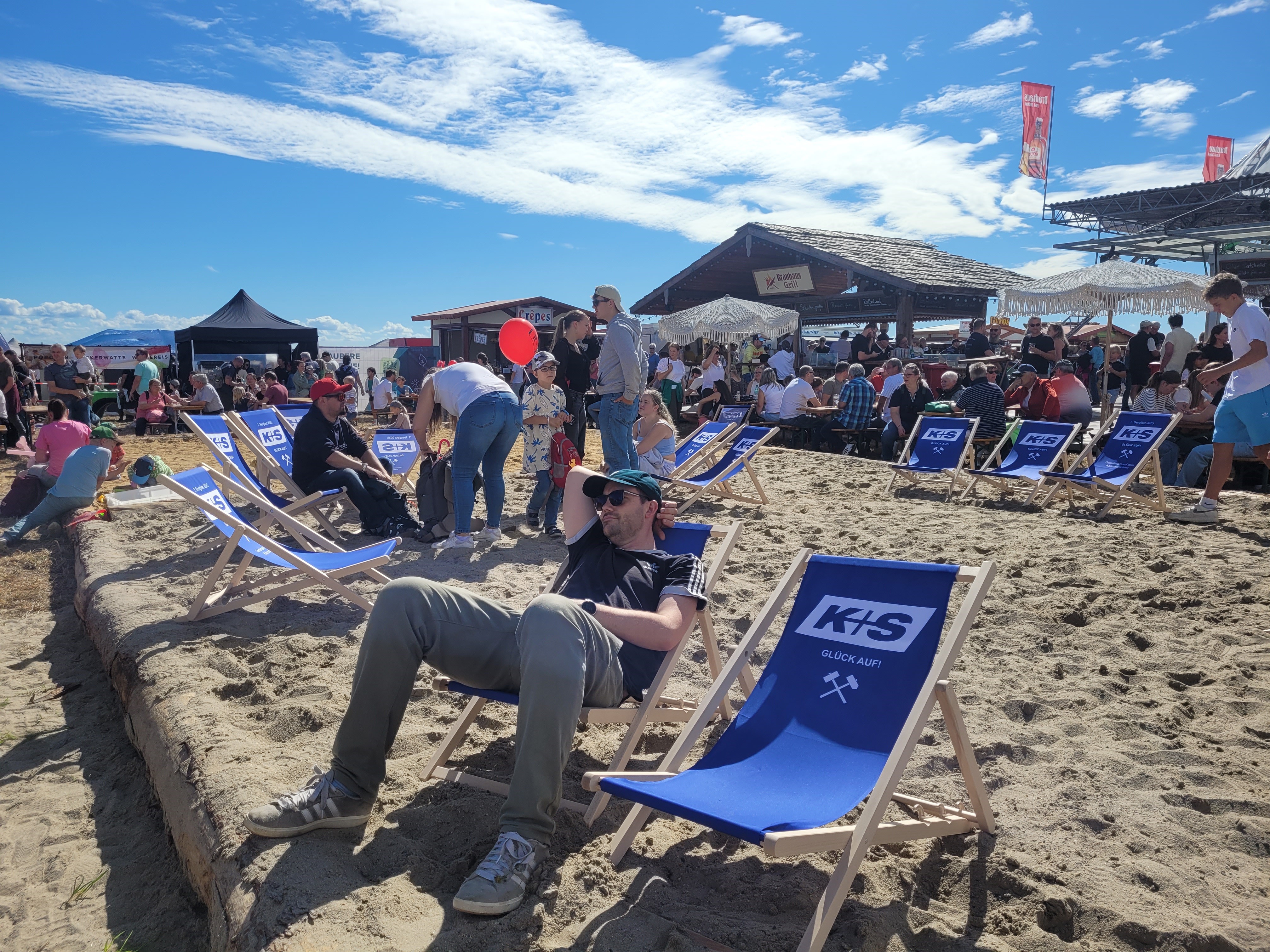 Das Bild zeigt einen sonnigen Tag auf einer Sandfläche, auf der viele Menschen auf blauen Liegestühlen mit der Aufschrift „K+S Glück Auf“ entspannen. Im Hintergrund sind Gebäude und Stände zu sehen sowie eine große Menschenmenge, die sich unterhält und das Wetter genießt. Einige Personen stehen, andere sitzen oder liegen. Der Himmel ist blau mit weißen Wolken.