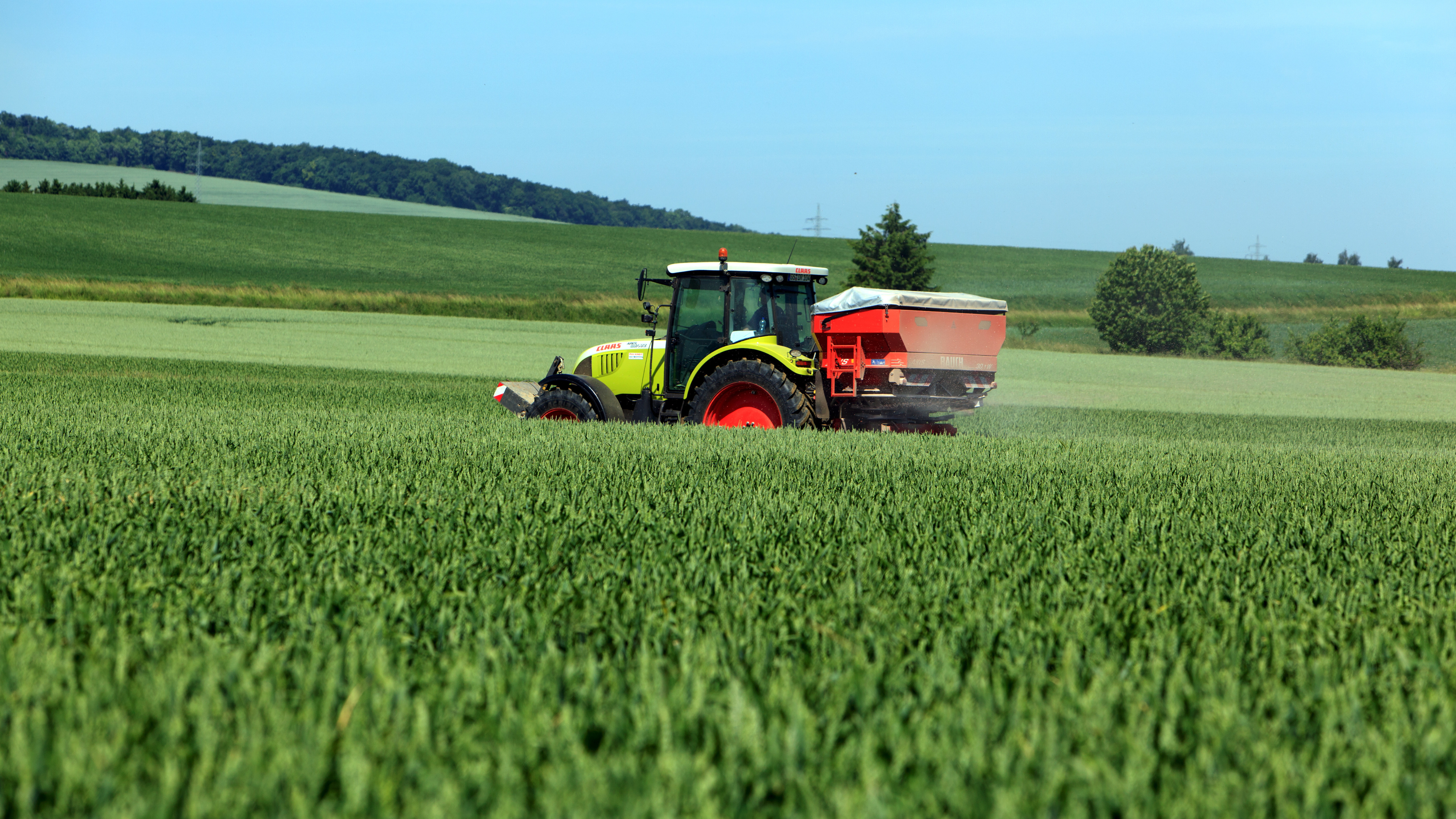 Das Bild zeigt einen Traktor, der auf einem großen, grünen landwirtschaftlichen Feld arbeitet. Am Heck ist ein roter Anbau angebracht, vermutlich zum Ausbringen von Dünger oder Saatgut. Im Hintergrund sind sanfte Hügel und Bäume unter einem klaren blauen Himmel zu sehen.