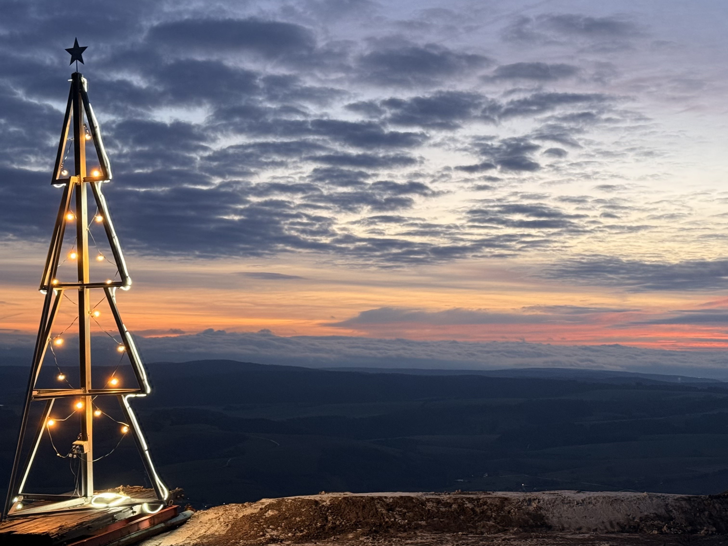 Metallkonstruktion in Form eines Weihnachtsbaums mit leuchtenden Lichtern und Stern an der Spitze, aufgestellt auf dem Plateau einer Salzhalde vor einem weiten Landschaftspanorama bei Sonnenaufgang mit farbigem Himmel und Wolken.