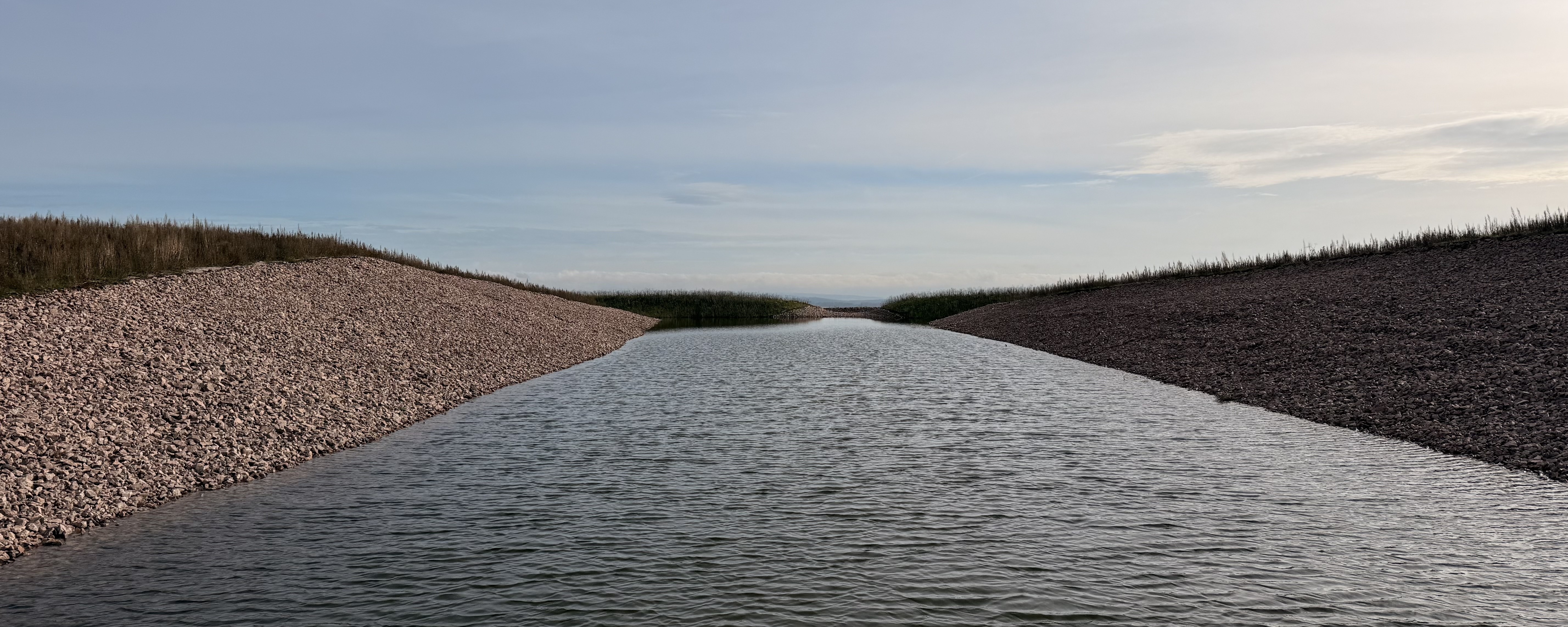 Retention basin on a vegetated tailings pile, featuring a water-filled basin and riprap-armored slopes under an overcast sky.