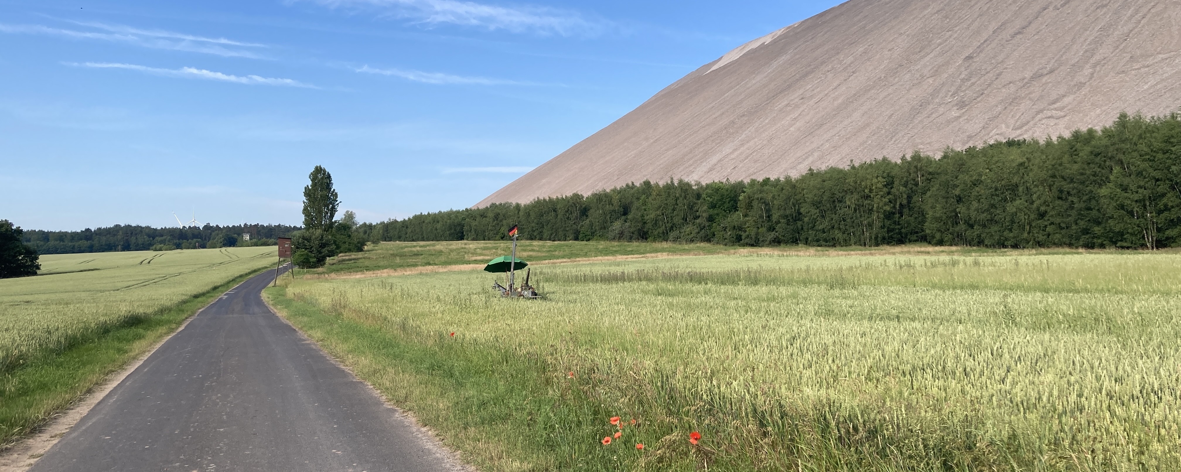 Kleinbohrgerät zur Untersuchung des Untergrunds auf einer Grünfläche am Fuß einer Halde. Im Hintergrund sind die Haldenflanke, Waldflächen und landwirtschaftlich genutzte Flächen zu sehen.