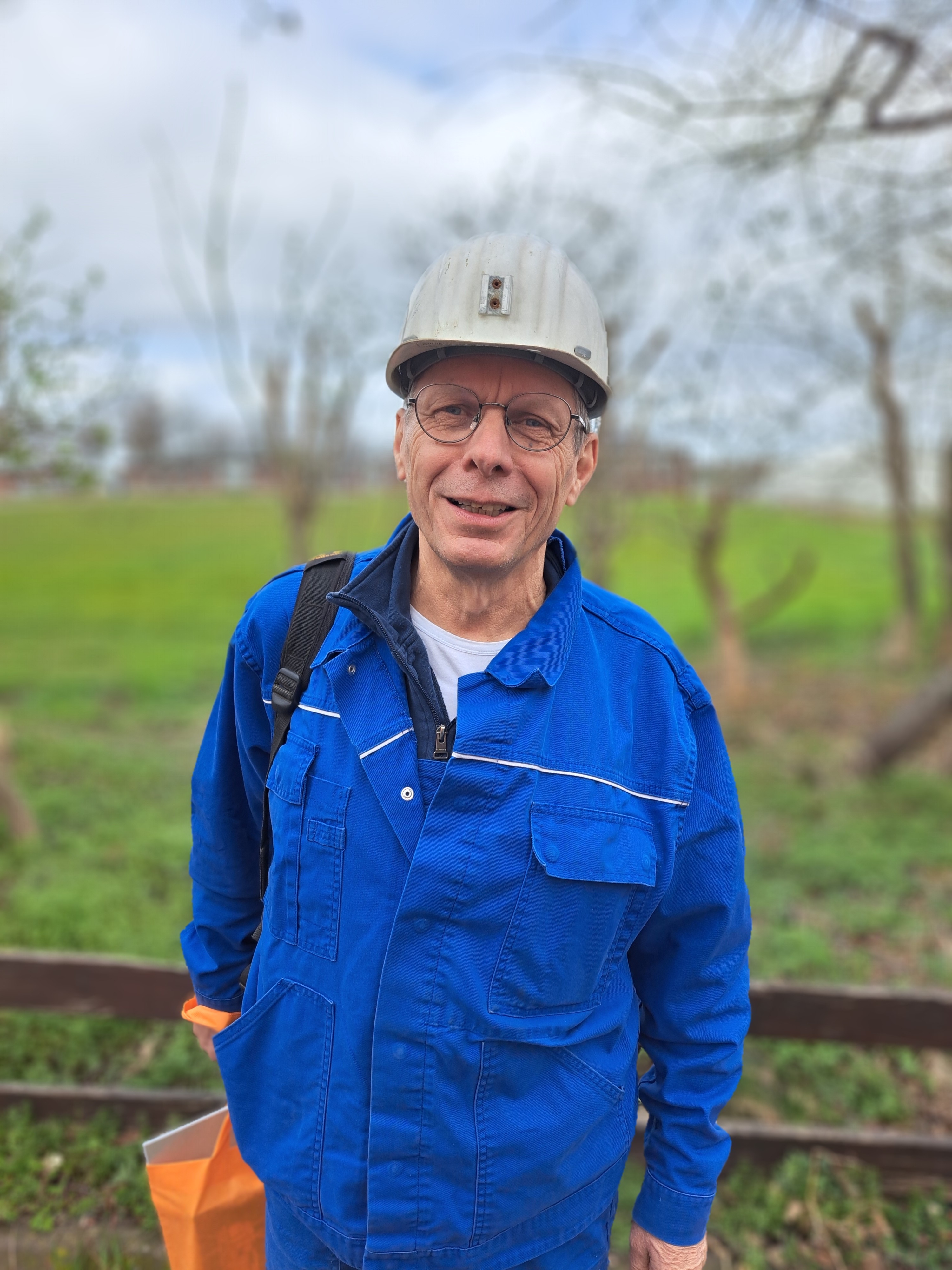 Eine Person in blauer Arbeitskleidung und weißem Schutzhelm steht im Freien. Die Person trägt eine orangefarbene Tasche und hat einen schwarzen Schultergurt. Im Hintergrund sind grüne Wiese, Bäume und ein bewölkter Himmel zu sehen.