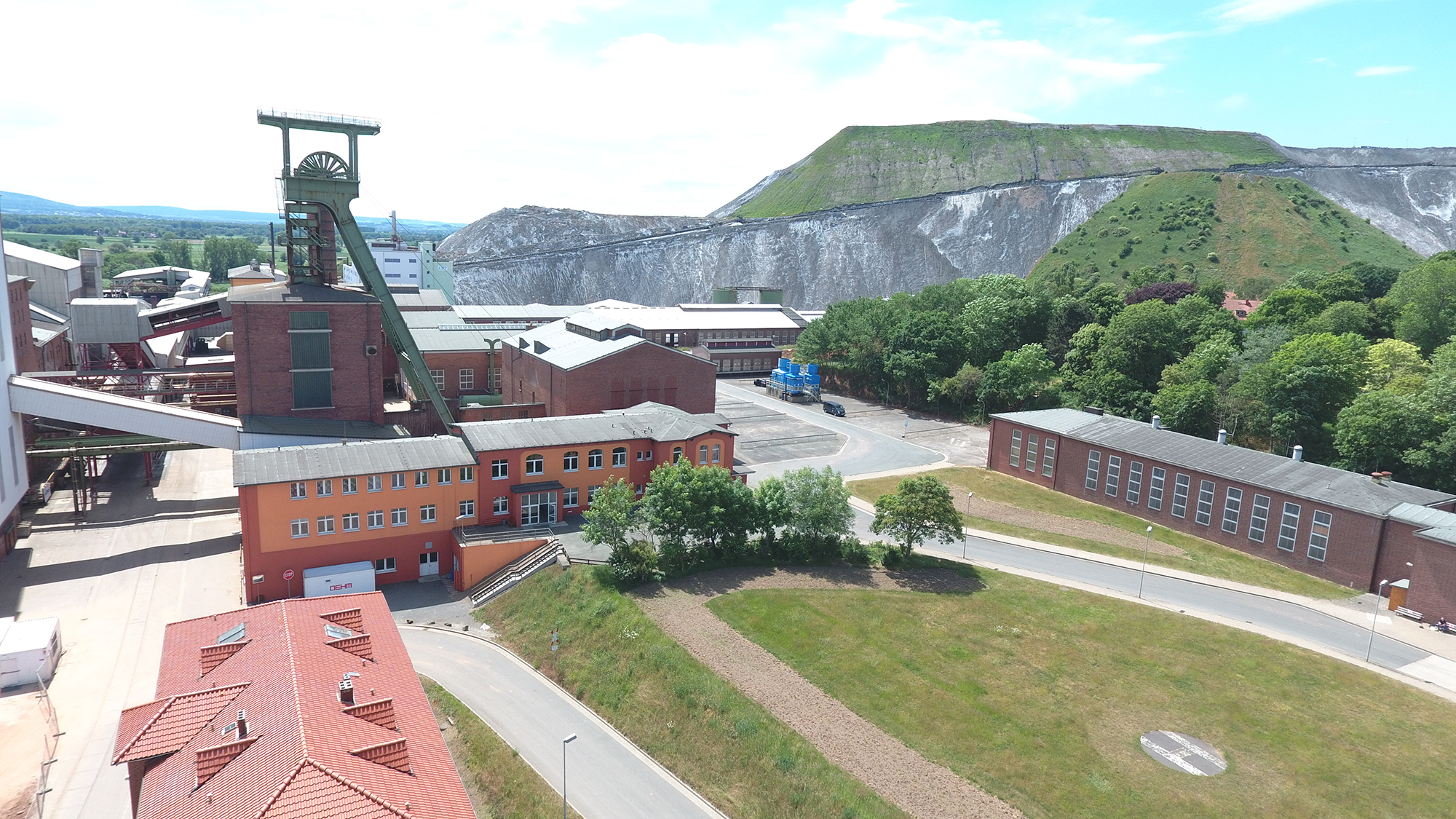 The image shows the Sigmundshall site with a headframe. Several buildings are visible, including a prominent brick building with an attached headframe structure. In the background, there is a large heap that is partially covered with vegetation.
