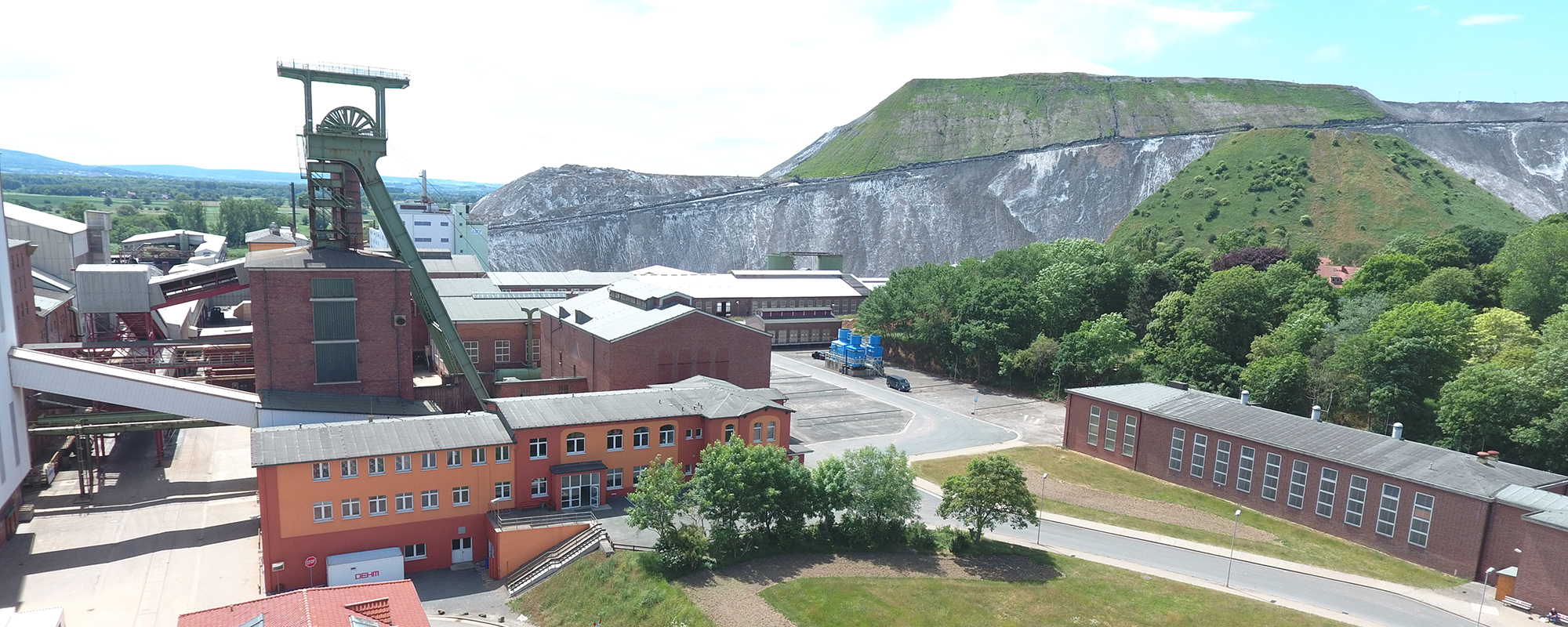 The image shows the Sigmundshall site with a headframe. Several buildings are visible, including a prominent brick building with an attached headframe structure. In the background, there is a large heap that is partially covered with vegetation.