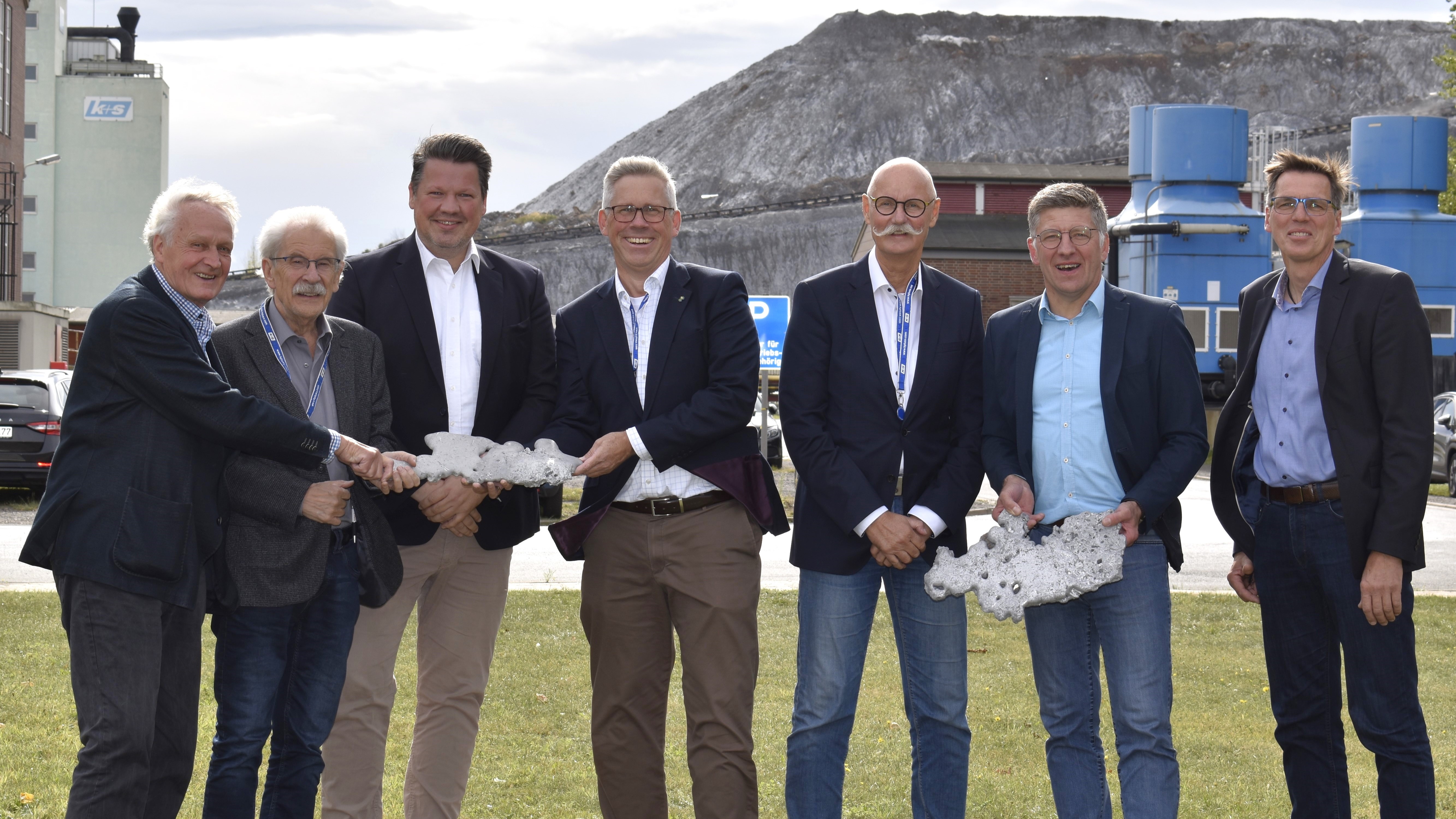 Eine Gruppe von sieben Personen steht auf einer grasbewachsenen Fläche vor einem industriellen Hintergrund mit Gebäuden und Maschinen. Die Personen tragen Businesskleidung, einige halten Gesteins- oder Mineralsamples in den Händen.