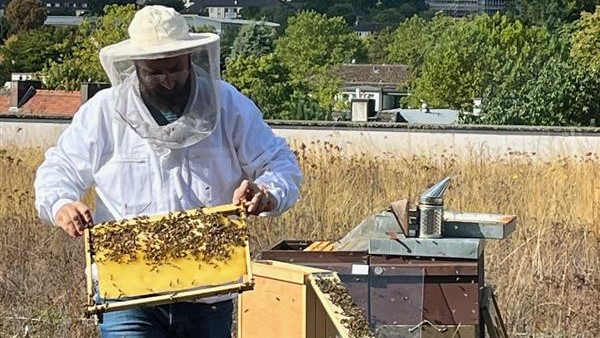 The Kassel city beekeeper stands on the roof of the K+S headquarters. The beehive is open and he is holding a honeycomb to show it to the children.