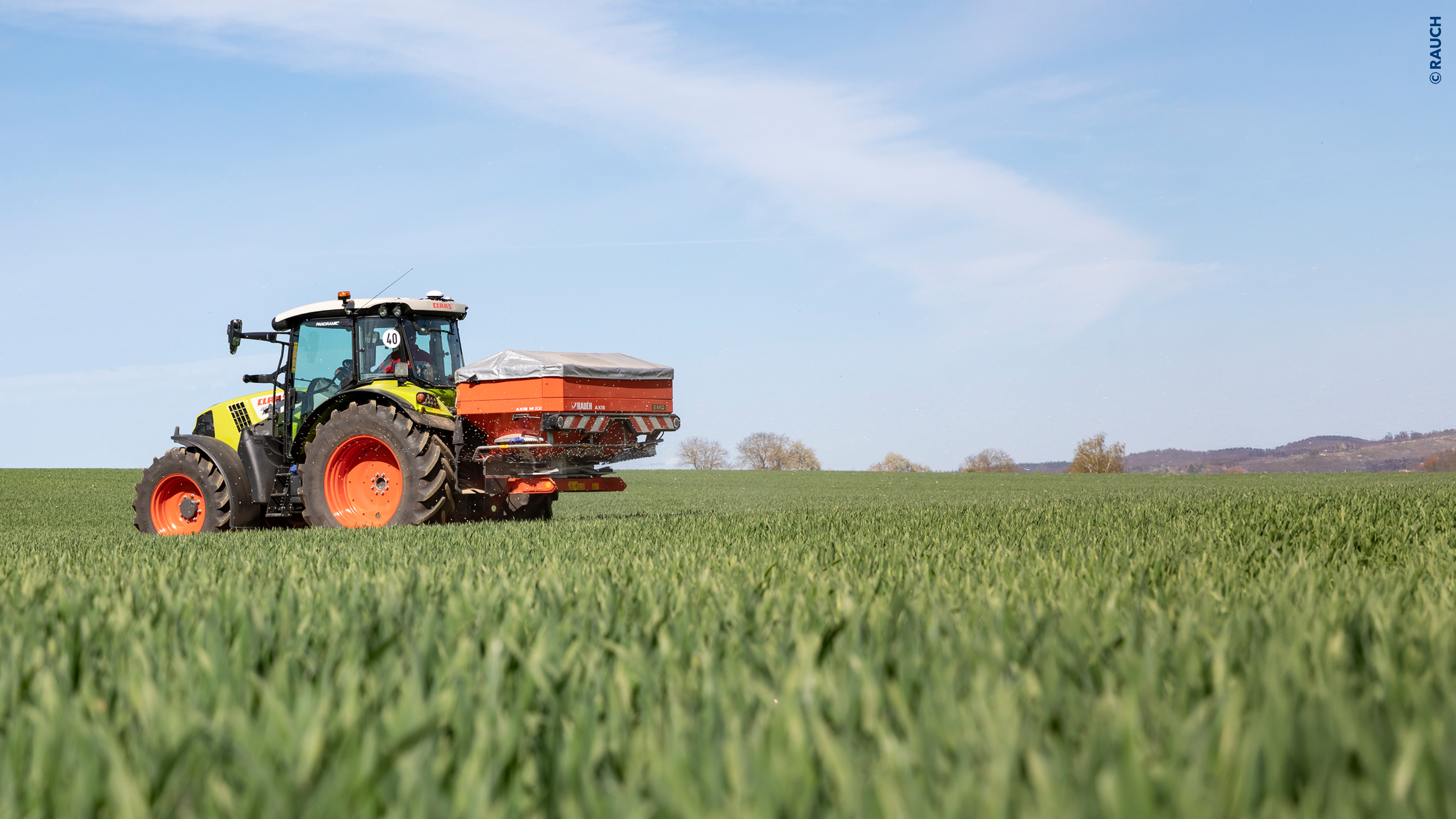 Traktor mit Düngerstreuer auf einem grünen Getreidefeld.
