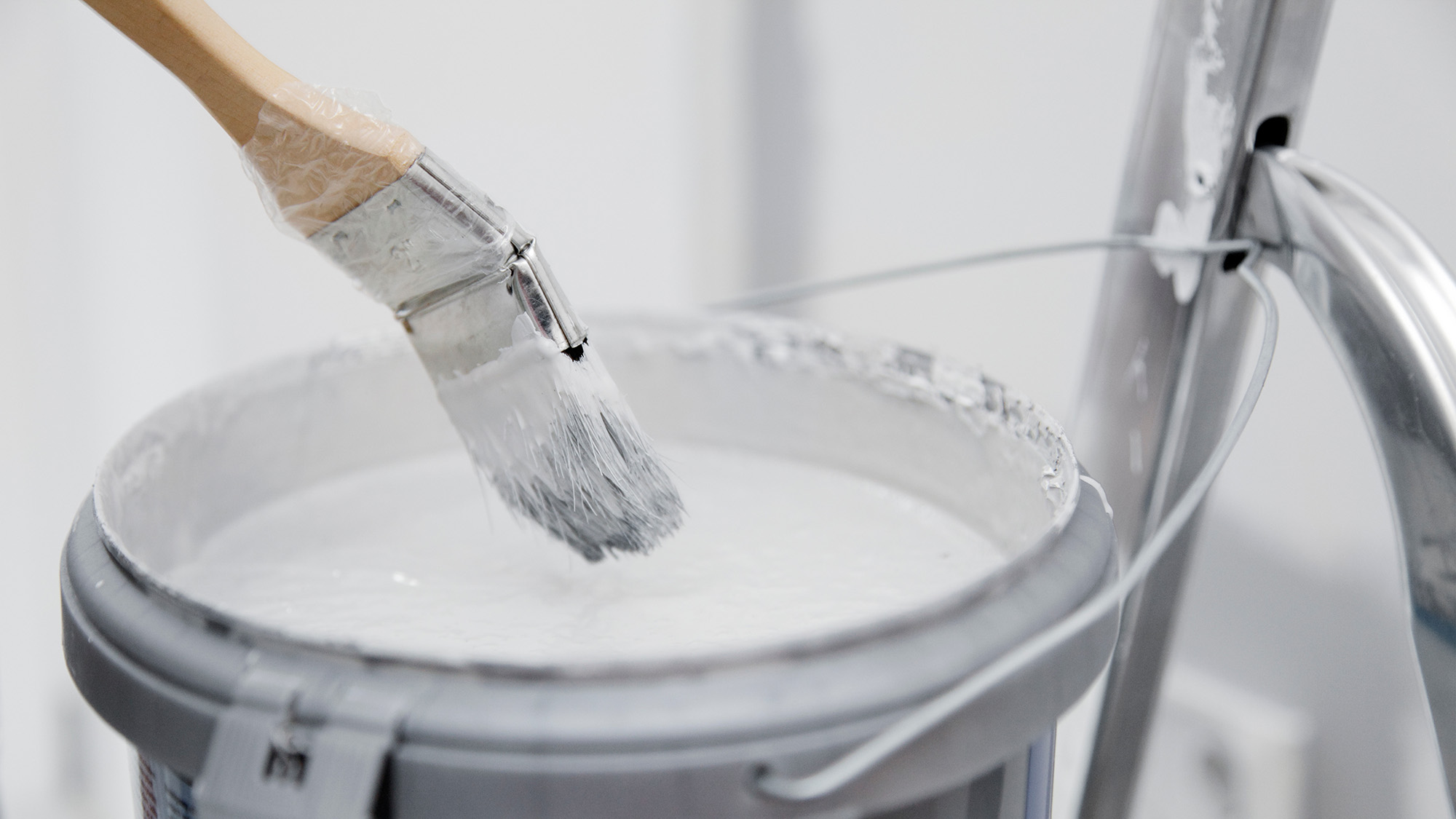 Pinsel ueber offenem Farbeimer mit weisser Farbe neben einem Metallgriff