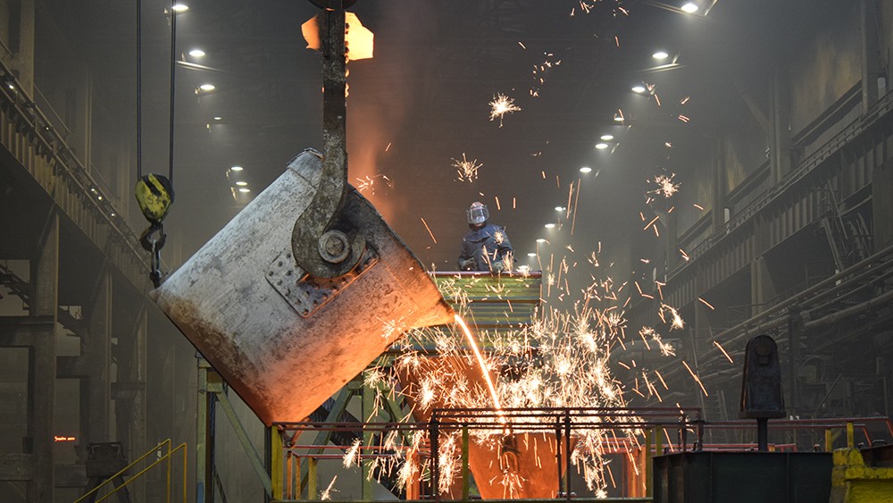 Industriehalle mit einem per Kran gekippten Giessbehaelter, aus dem gluessendes Metall in einen grossen Behälter fliesst; Funken spruehen, im Hintergrund steht eine Person auf einer Plattform.