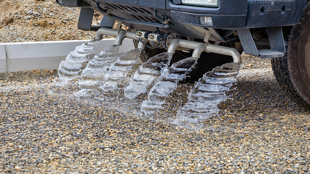 Underside of a vehicle with several pipes spraying wide streams of water onto a gravel surface.