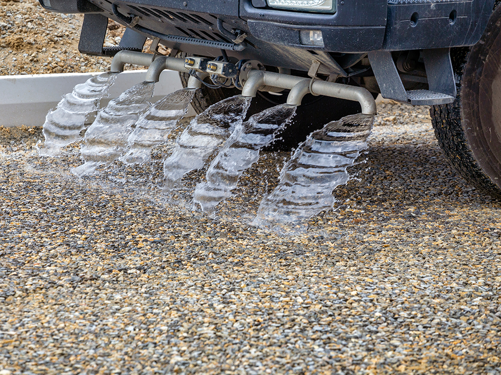 Underside of a vehicle with several pipes spraying wide streams of water onto a gravel surface.