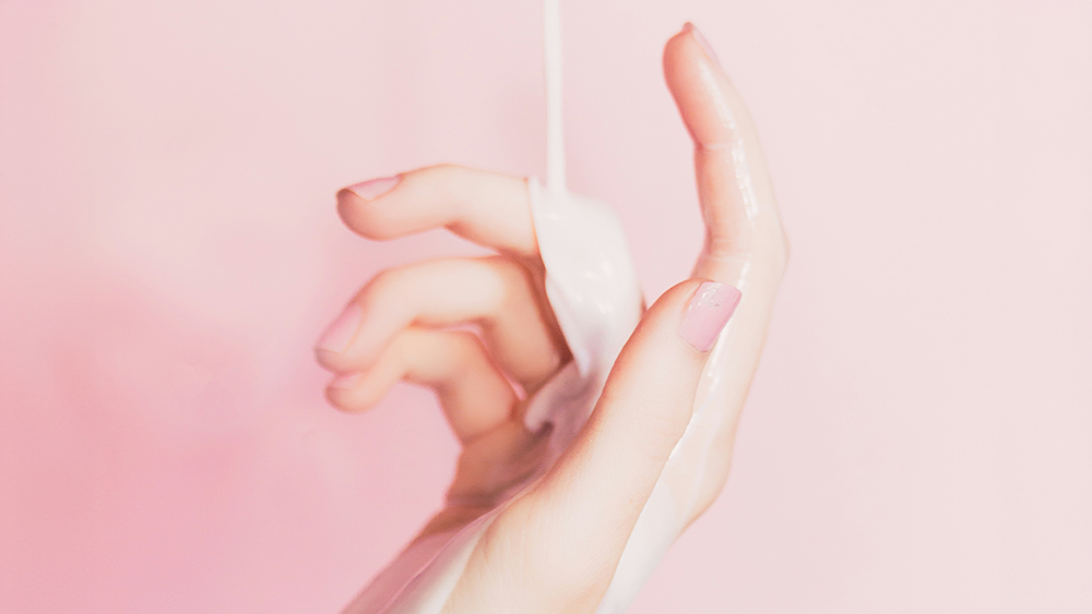 Nahaufnahme einer Hand, die ein weisses Wattepad vor einem zartrosa Hintergrund haelt.
