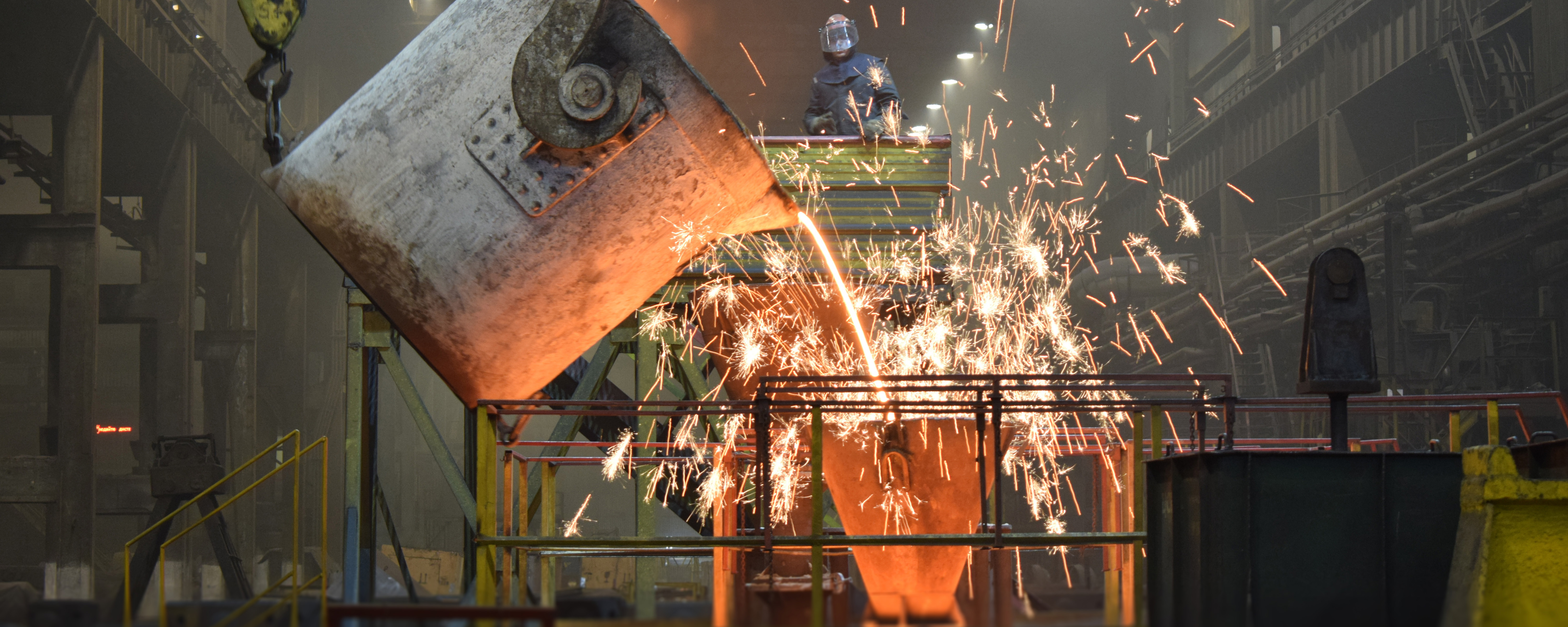 Flüssiges Metall wird in einer Industriehalle aus einem großen Kessel gegossen, während Funken in alle Richtungen sprühen.