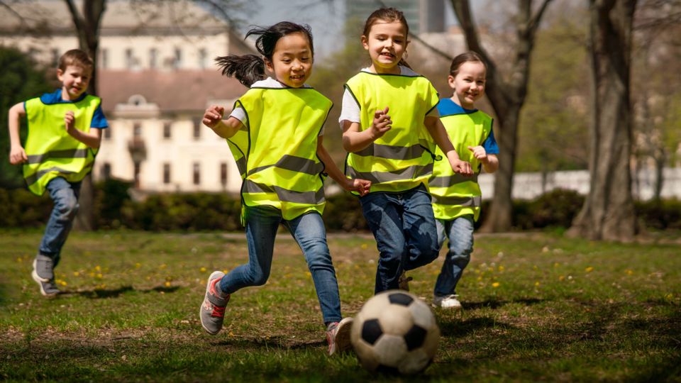 Vier Kinder spielen Fußball im Freien. Sie tragen gelbe Sicherheitswesten. Im Hintergrund sind Bäume und ein Gebäude zu sehen.