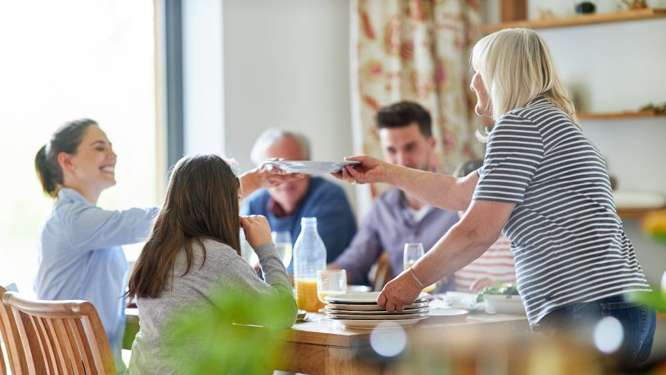Familie sitzt am Esstisch für eine gemeinsame Mahlzeit und reicht sich strahlende Teller